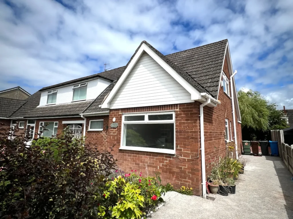 A brick house with a triangular roof section, a large front window, and a small garden with various plants. A pathway leads to the side of the house under a partly cloudy blue sky.